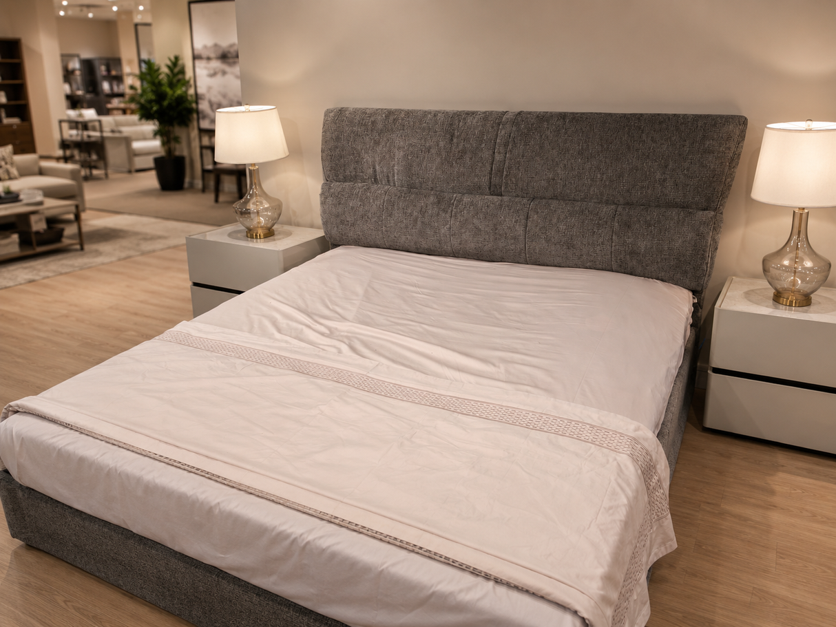 A modern bed with a tall, tufted gray upholstered headboard and matching base, dressed in white bedding and a light beige throw blanket, set in a contemporary bedroom showroom with two bedside tables and lamps.