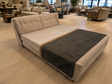 A modern upholstered daybed or sofa bed in light beige fabric, shown in a furniture showroom with light wood flooring and other seating pieces in the background. A dark charcoal gray throw blanket is draped across the front.