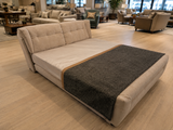 A modern upholstered daybed or sofa bed in light beige fabric, shown in a furniture showroom with light wood flooring and other seating pieces in the background. A dark charcoal gray throw blanket is draped across the front.