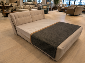 A modern upholstered daybed or sofa bed in light beige fabric, shown in a furniture showroom with light wood flooring and other seating pieces in the background. A dark charcoal gray throw blanket is draped across the front.