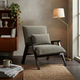 A modern upholstered accent chair in a sage green fabric with dark wood arms and legs, styled in a cozy room with a matching pillow, a floor lamp, a bookshelf, and a rug.