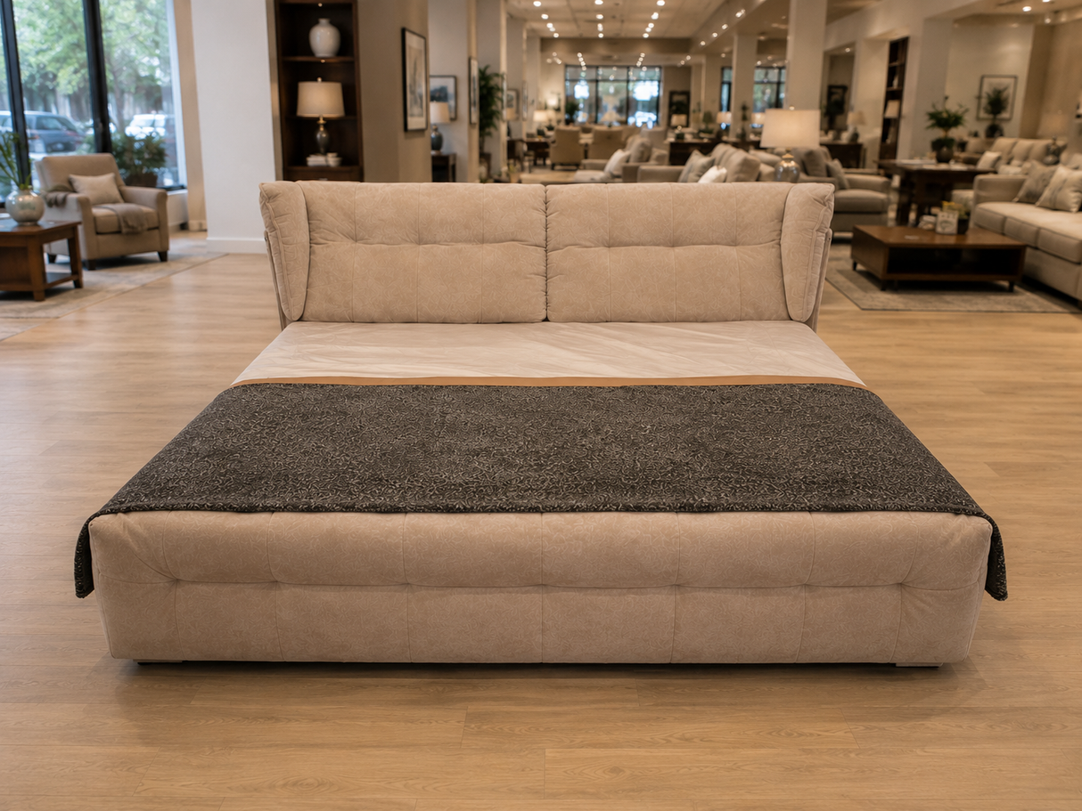 A modern upholstered platform bed with a high tufted headboard, shown in a furniture showroom. The bed is upholstered in a light beige fabric and features a dark charcoal gray textured throw blanket draped across the foot.
