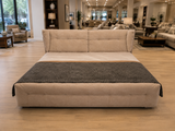 A modern upholstered platform bed with a high tufted headboard, shown in a furniture showroom. The bed is upholstered in a light beige fabric and features a dark charcoal gray textured throw blanket draped across the foot.