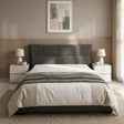 A modern bed with a dark gray upholstered headboard, dressed in white and beige bedding, set in a minimalist bedroom with a slatted wood headboard panel, bedside tables, lamps, and a textured rug.